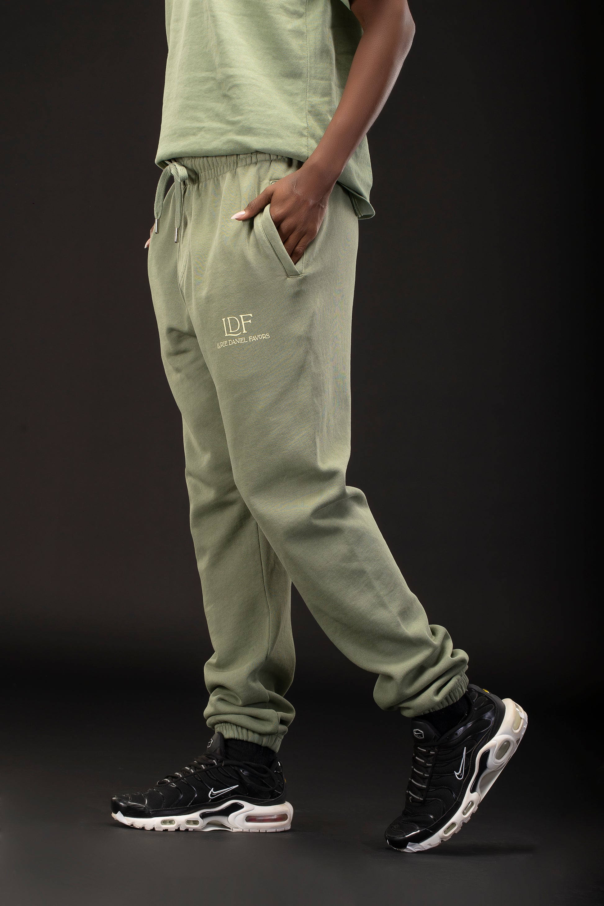 Person wearing green jogger pants with a logo and black sneakers on a dark background
