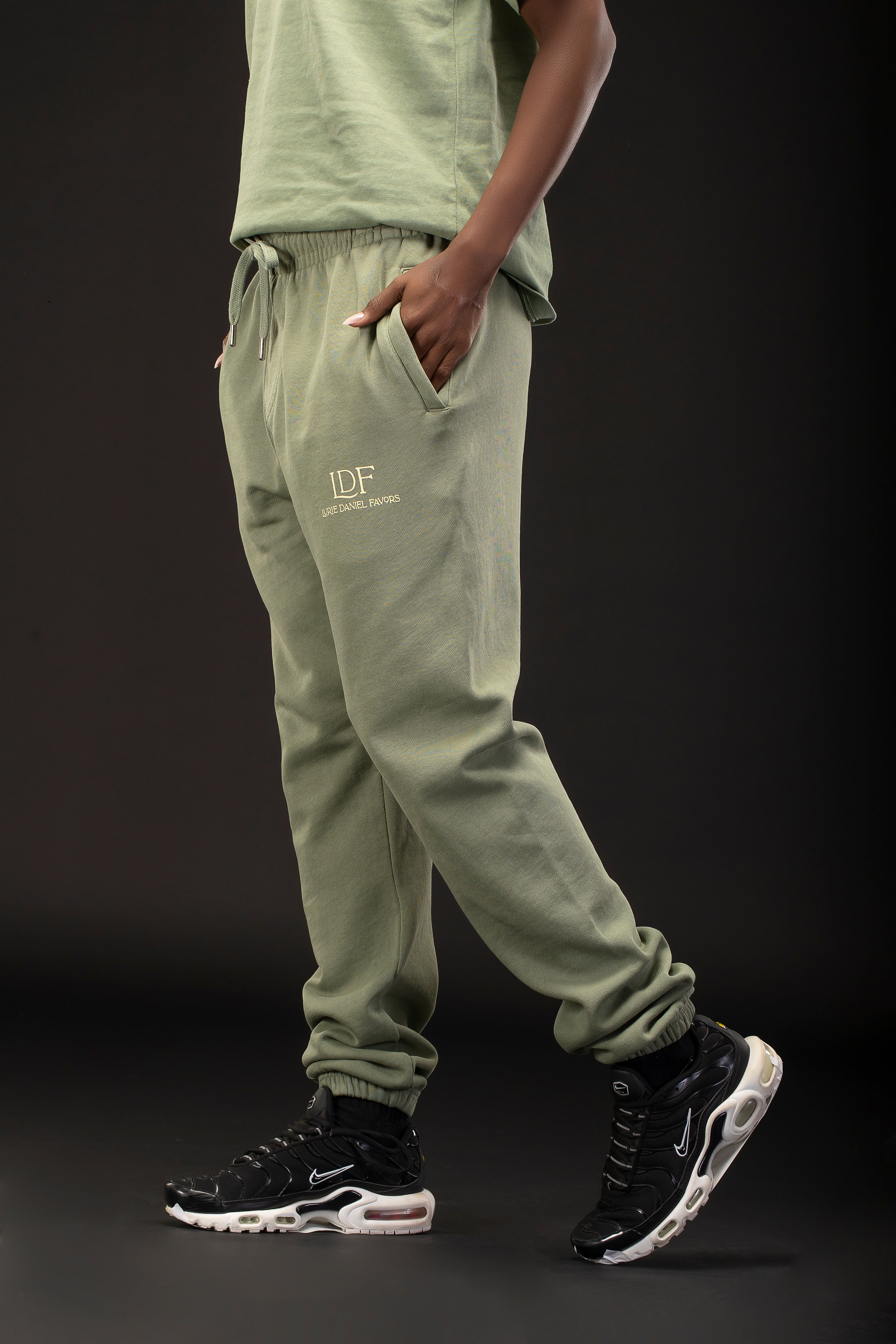 Person wearing green jogger pants with a logo and black sneakers on a dark background
