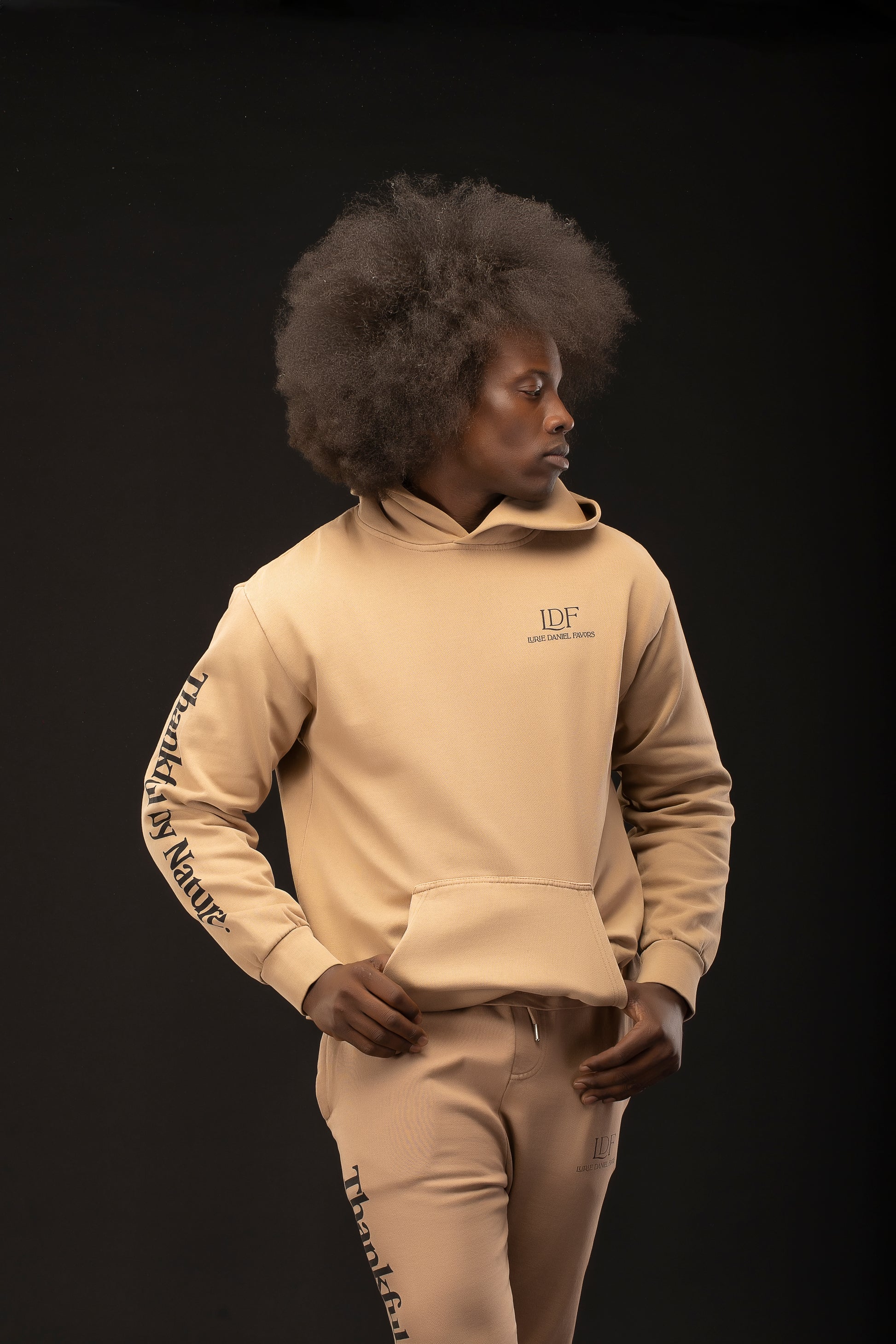 Man wearing a beige hoodie and pants with visible branding on a black background