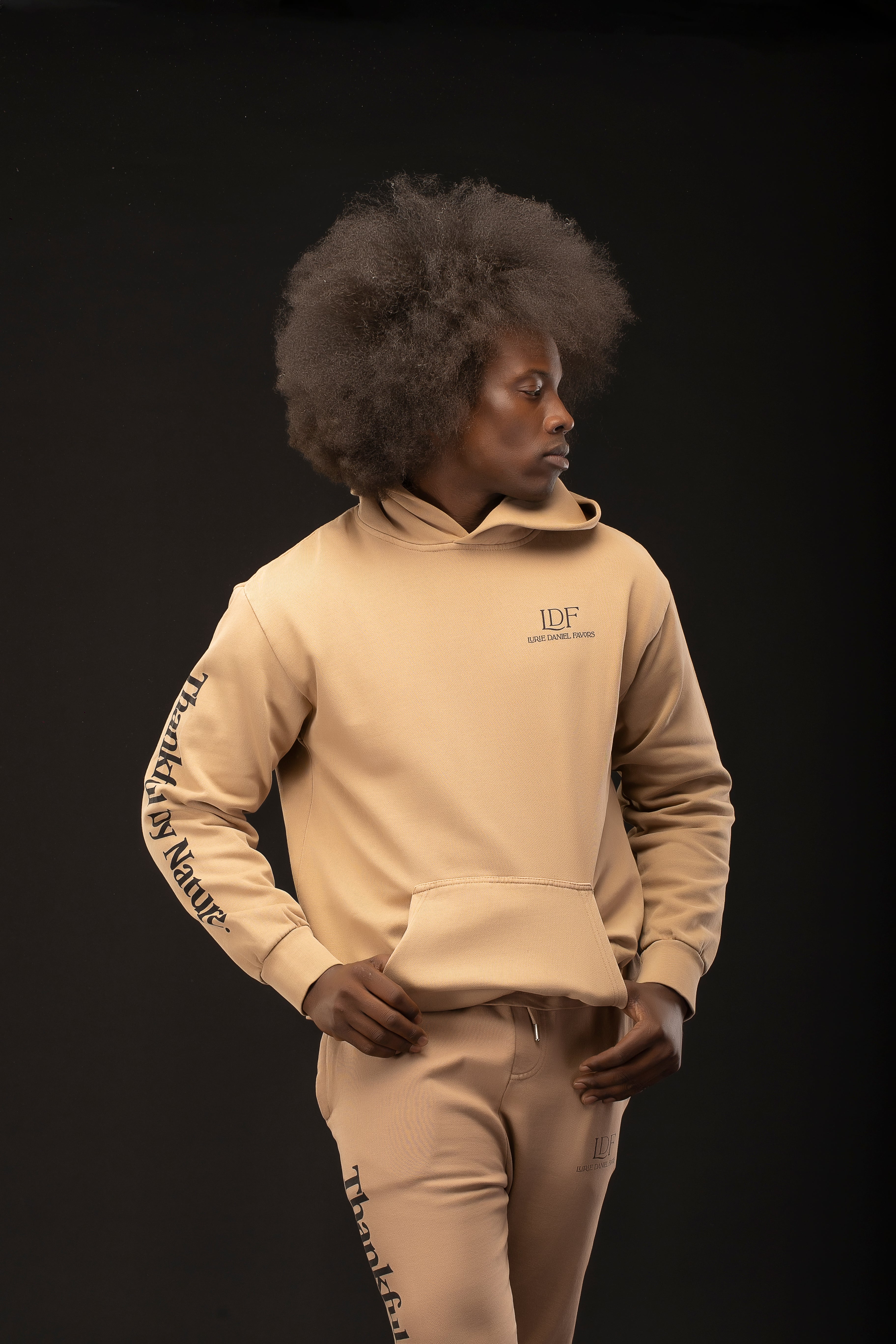 Man wearing a beige hoodie and pants with visible branding on a black background