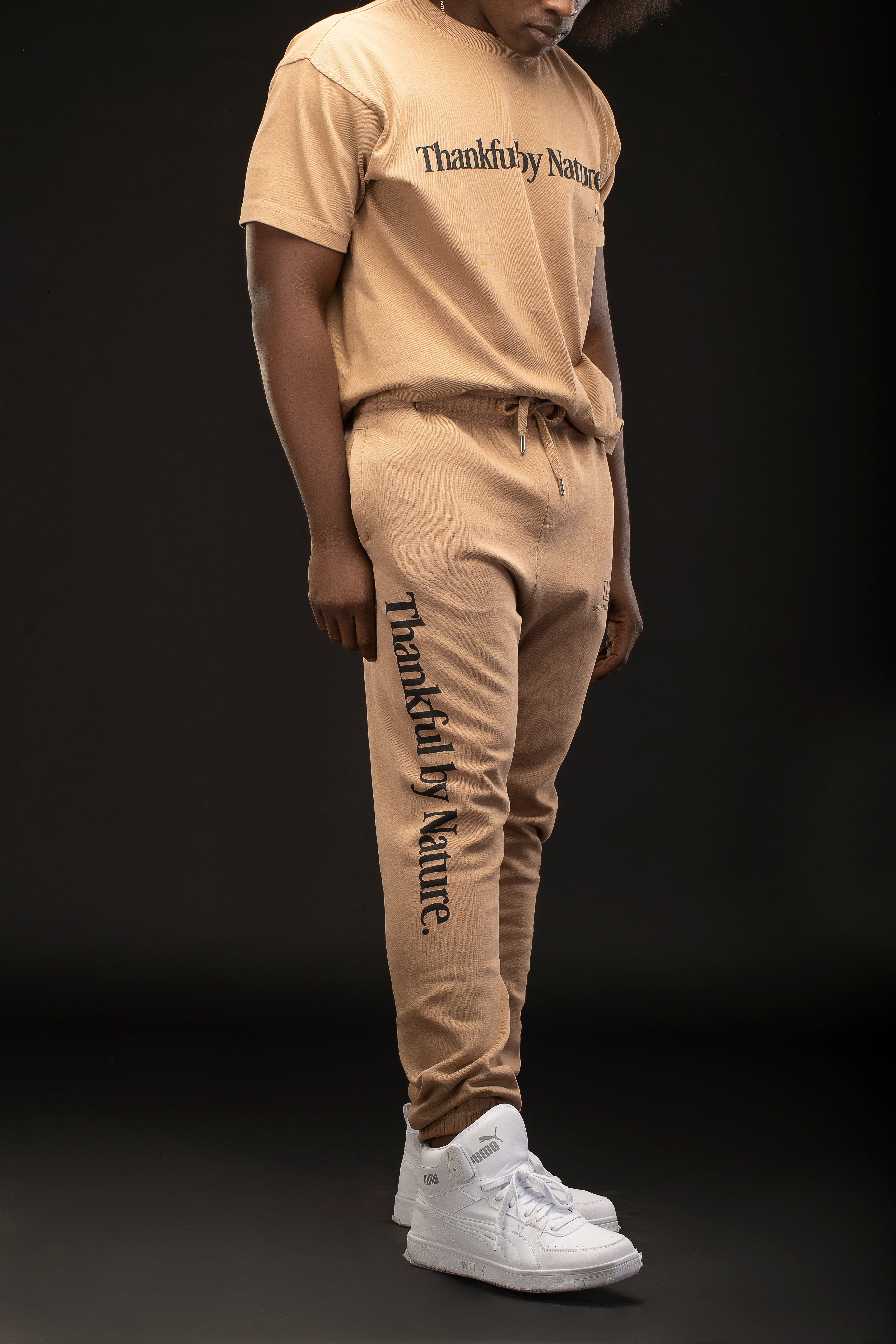 Man wearing a beige 'Thankful by Nature' outfit with white sneakers on a black background